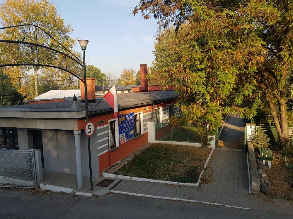 Budynek z numerem 5, pomalowany na szaro i pomarańczowo, z polską flagą na dachu, otoczony zielenią i chodnikiem z kostki brukowej.