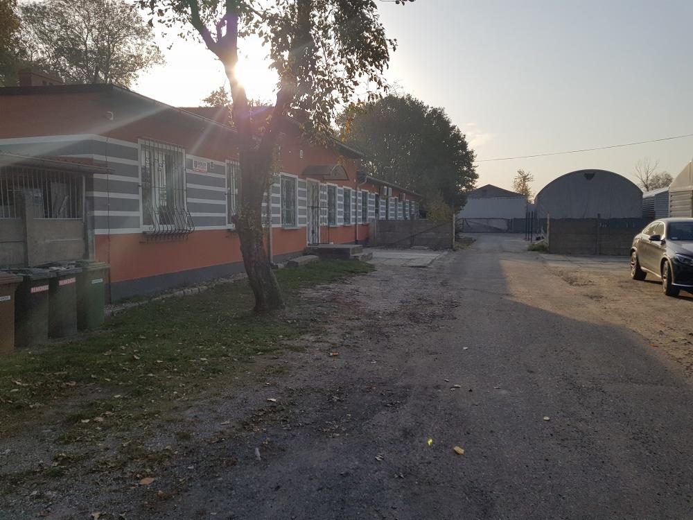 Widok na budynek z pomarańczową elewacją, szare pasy pod oknami i metalowe kraty w oknach, z widoczną drogą dojazdową, samochodem po prawej i konstrukcjami namiotowymi w tle, w słoneczny dzień.