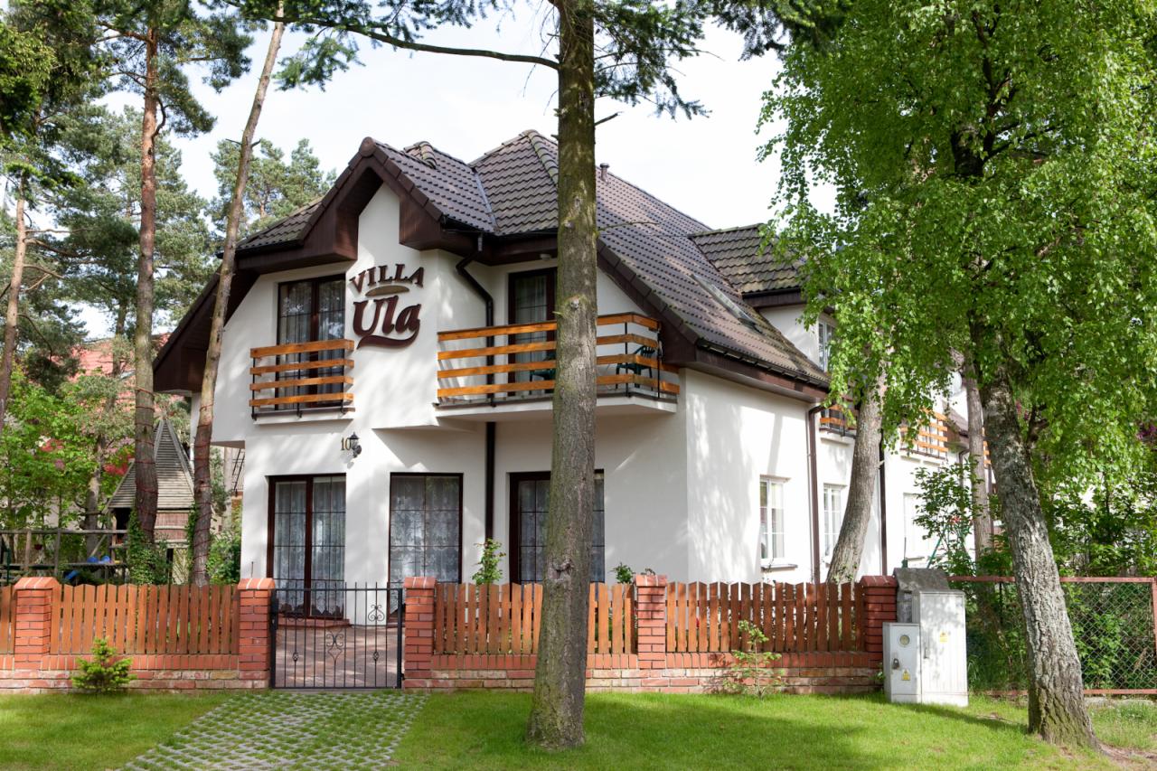 Dwupiętrowa willa z brązowym dachem i balkonami, otoczona zielenią, z widocznym napisem 'Villa Ula' nad balkonem.