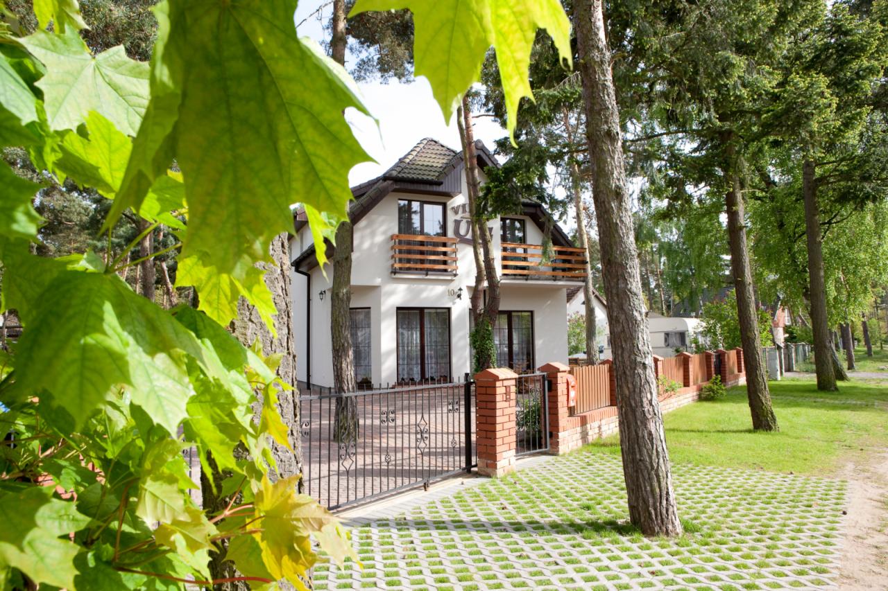 Biały budynek z brązowymi balkonami, widoczny przez drzewa i liście klonu, z czarnym ogrodzeniem i ażurową kostką brukową.