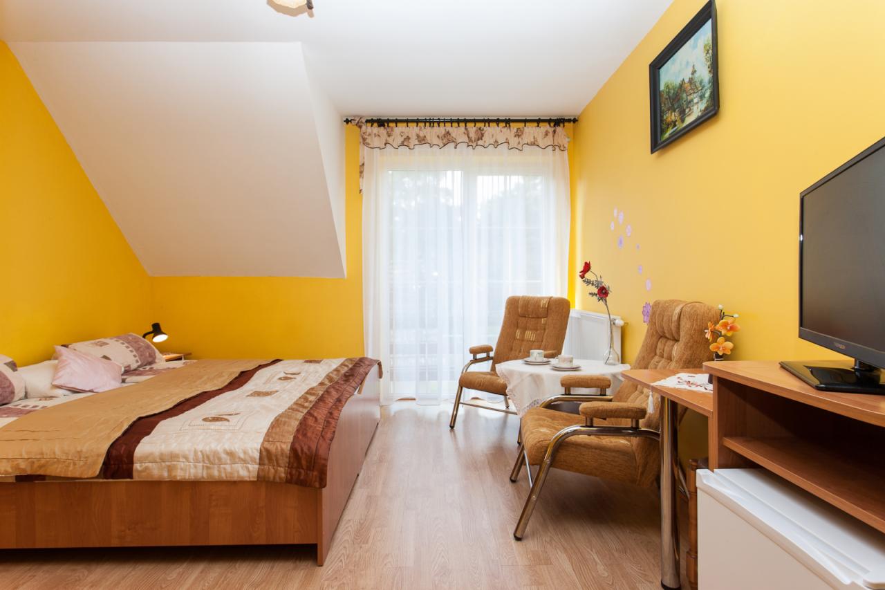 Pokój w pensjonacie w Pobierowie z żółtymi ścianami, łóżkiem z brązową narzutą, dwoma fotelami ze stoliczkiem i telewizorem na drewnianej komodzie.