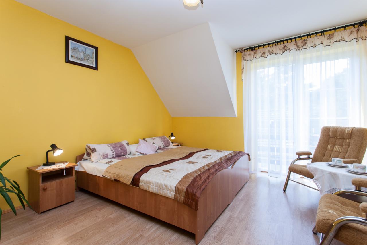 Pokój hotelowy z żółtymi ścianami, drewnianym łóżkiem z wzorzystą narzutą, stolikiem nocnym z lampką, dwoma fotelami przy stoliku nakrytym białym obrusem i oknem z firankami.