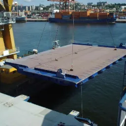 Żuraw portowy podnosi stalową platformę nad wodą, w tle widoczne statki i zabudowa portowa.