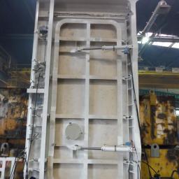 Vogen sp. z o.o. - Wysoka, biała, kratownicowa konstrukcja stalowa z siłownikami hydraulicznymi, zawieszona na haku suwnicy w hali produkcyjnej.