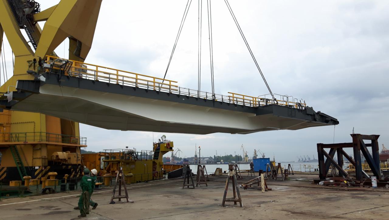 Żółty dźwig portowy podnosi stalową konstrukcję mostu z żółtymi barierkami, widoczni dwaj pracownicy w zielonych kombinezonach, w tle port z żurawiami.