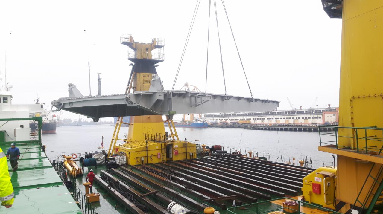Transport i montaż dużej stalowej konstrukcji dźwigiem na pokładzie statku w porcie, widoczne nabrzeże i budynki portowe w tle.