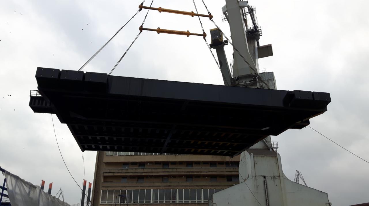 Ciężki, czarny element konstrukcyjny podnoszony przez dźwig na tle budynku i pochmurnego nieba. Widoczne liny i haki dźwigu.