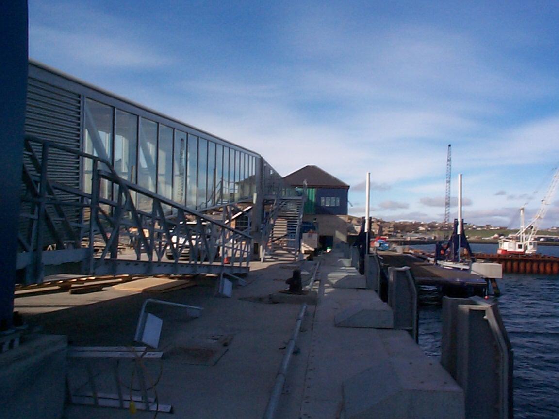 Metalowy pomost z przeszkloną konstrukcją nad wodą, widoczne dźwigi i nabrzeże w oddali.