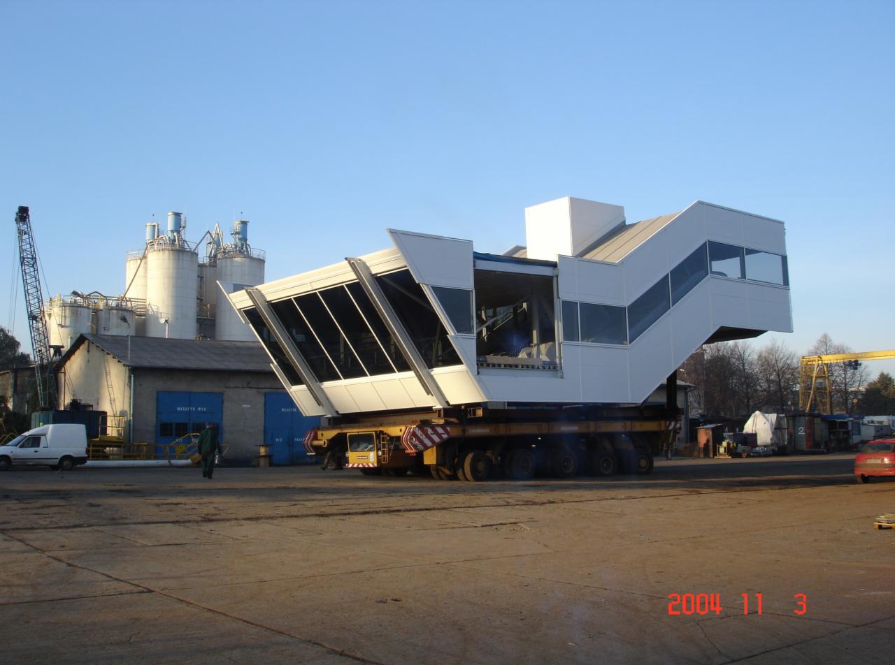 Transport modułowej konstrukcji stalowej z przeszklonymi ścianami na platformie transportowej, w tle budynki przemysłowe i silosy.