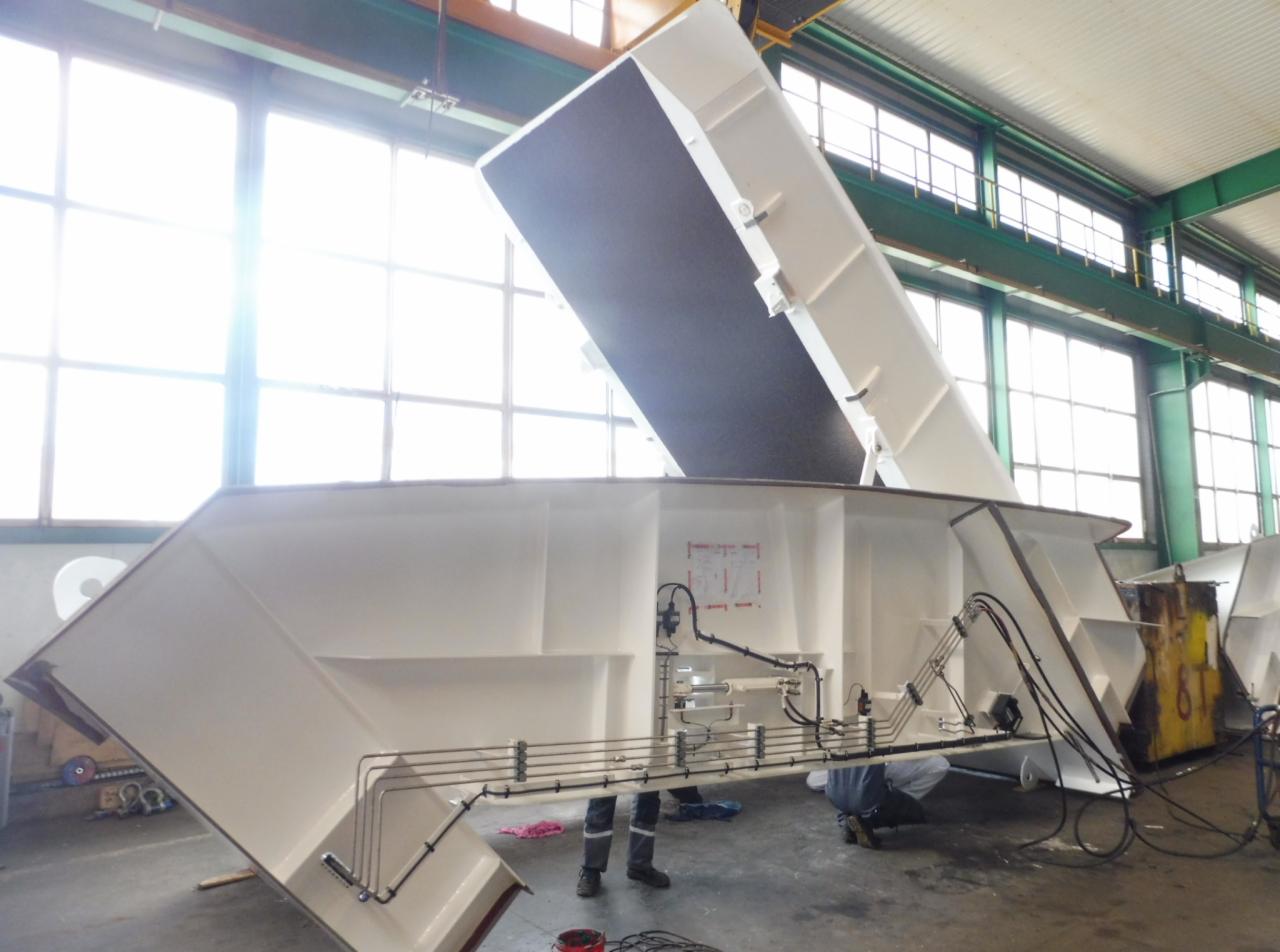 Wnętrze hali produkcyjnej z widoczną białą, metalową konstrukcją w trakcie montażu, z widocznymi elementami hydraulicznymi i schematem technicznym na ścianie konstrukcji.