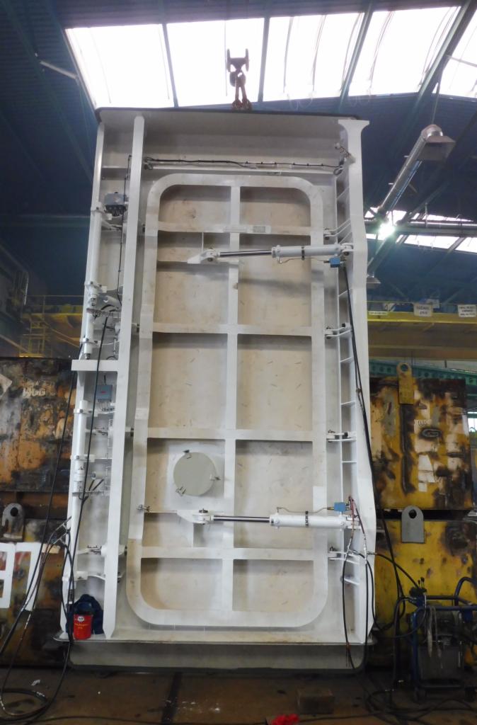 Wysoka, biała, kratownicowa konstrukcja stalowa z siłownikami hydraulicznymi, zawieszona na haku suwnicy w hali produkcyjnej.