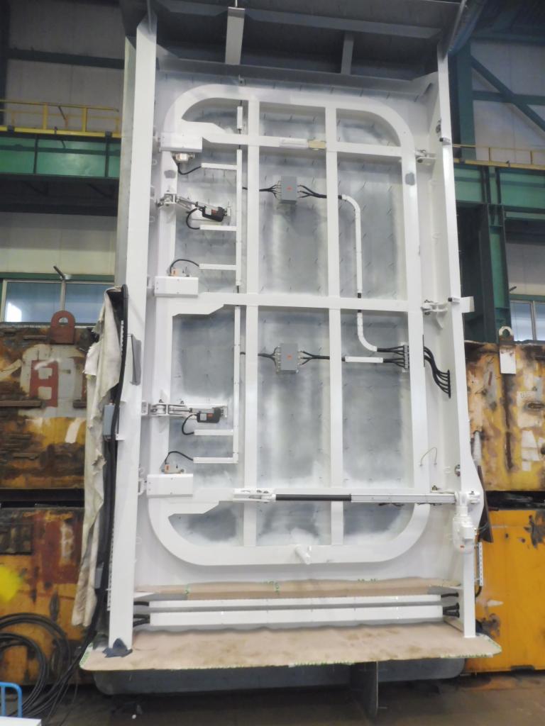 Wysoka, biała konstrukcja stalowa z elementami automatyki i okablowaniem, prawdopodobnie rama do hali lub specjalistycznego kontenera, widoczna w hali produkcyjnej z elementami rusztowań i zużytego...