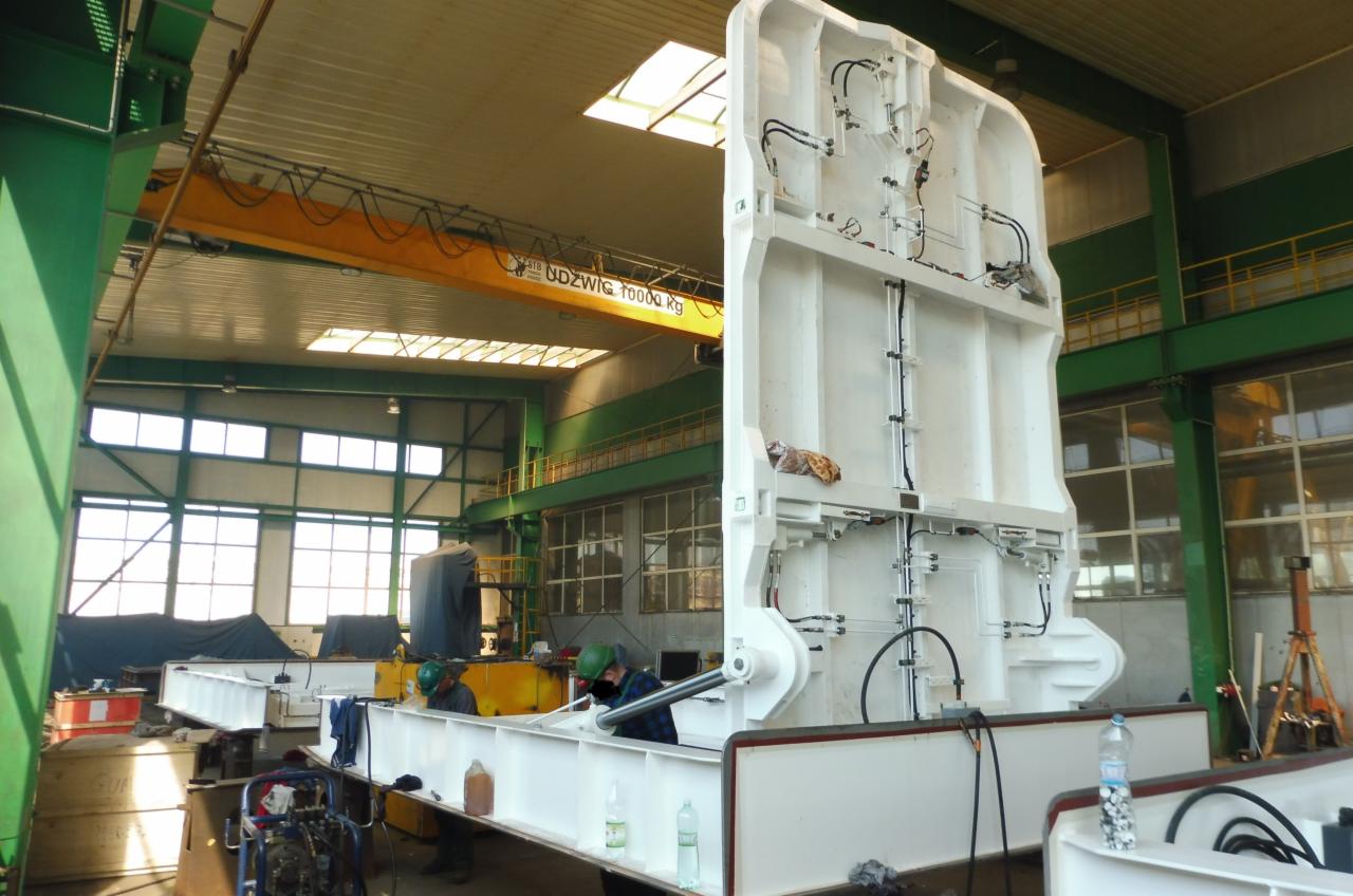 Wnętrze hali produkcyjnej z fragmentami spawanej, białej konstrukcji stalowej w trakcie montażu, widoczne okna, suwnica i pracownicy w kaskach.