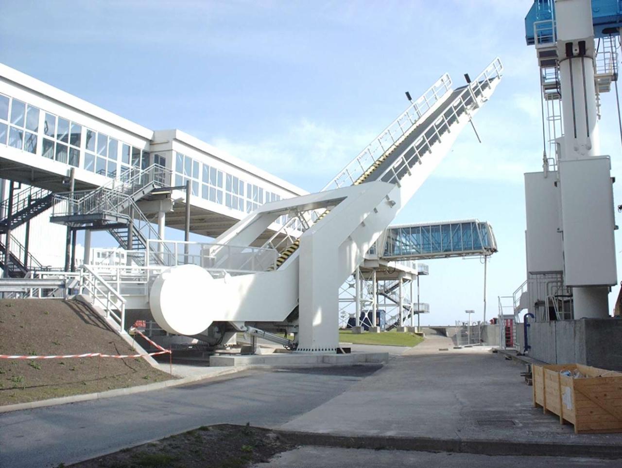 Biała, stalowa konstrukcja z ruchomymi schodami, prawdopodobnie przejście dla pasażerów w porcie lotniczym lub morskim, połączona z budynkiem za pomocą przeszklonego korytarza, widoczna skrzynia...