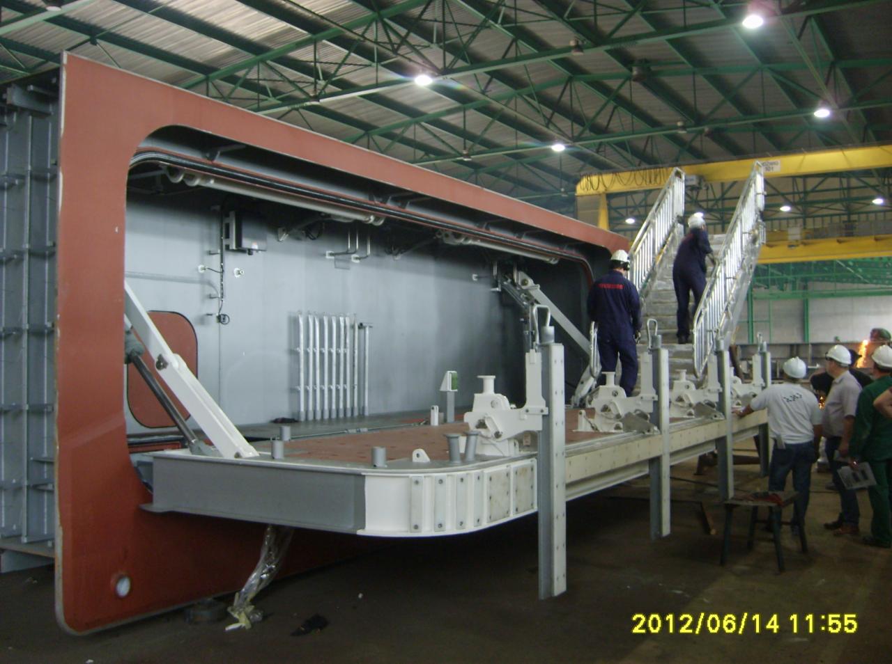 Wnętrze hali produkcyjnej z dużą, częściowo zmontowaną konstrukcją stalową, prawdopodobnie fragmentem statku, z pracownikami w kaskach na schodach w tle.