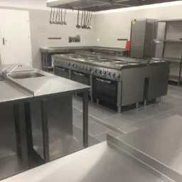 Profesjonalna kuchnia gastronomiczna ze stali nierdzewnej, widoczny piec z wieloma palnikami, zlewozmywaki, półki i wiszące przybory kuchenne.