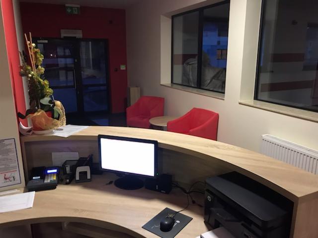 Recepcja warsztatu samochodowego z jasnym drewnianym biurkiem, komputerem, drukarką, czytnikiem kart i dwoma czerwonymi fotelami w tle.