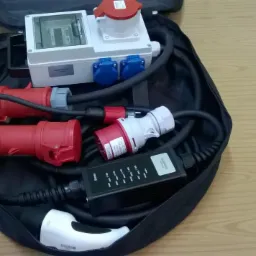 Zestaw przenośnych gniazd i wtyczek elektrycznych, w tym gniazda siłowe i adapter do ładowania pojazdów elektrycznych, umieszczony w czarnej torbie na drewnianym stole.