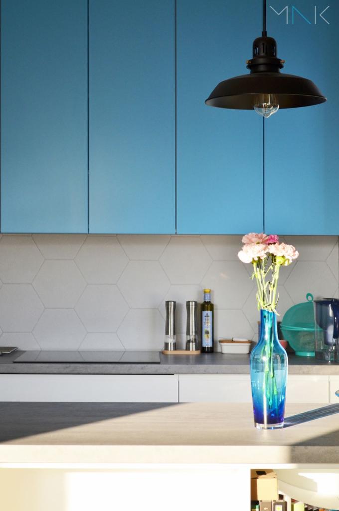 Nowoczesna kuchnia w Toruniu z błękitnymi szafkami, heksagonalnymi płytkami na ścianie i czarną lampą wiszącą, akcentowana niebieskim wazonem z różowymi kwiatami.