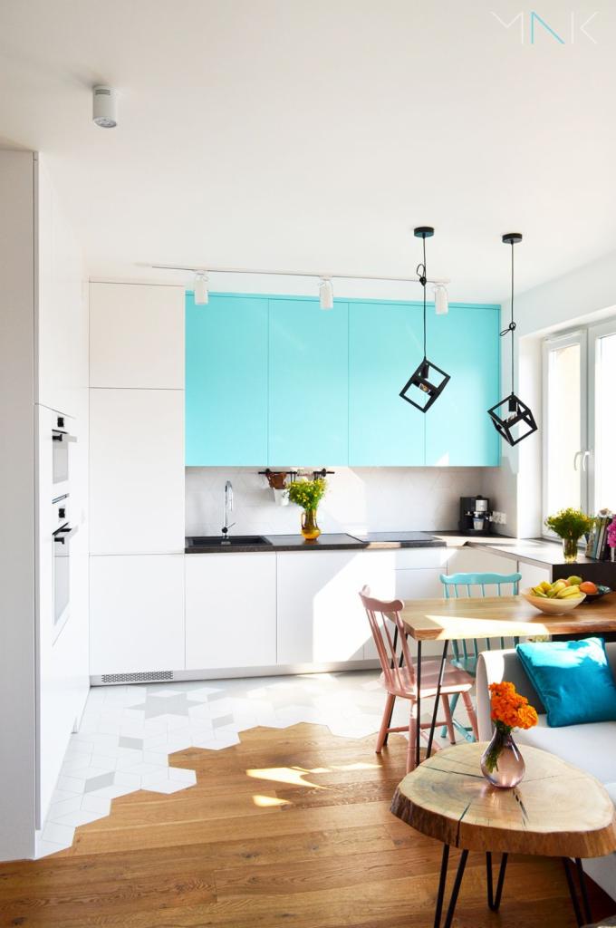 Jasne wnętrze kuchni z turkusowymi szafkami, drewnianą podłogą przechodzącą w heksagonalne płytki, drewnianym stołem i lampami w kształcie czarnych sześcianów.