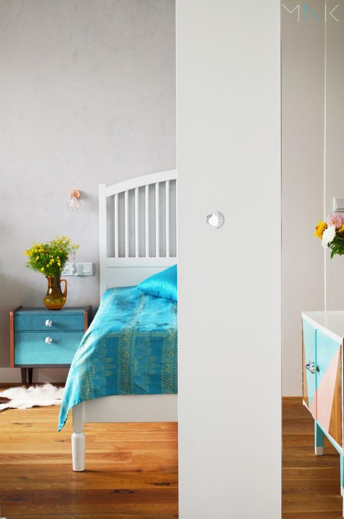 Aranżacja sypialni z turkusową pościelą na białym łóżku, drewnianą podłogą i kolorową szafką nocną w stylu retro, widziana przez częściowo otwarte białe drzwi przesuwne.