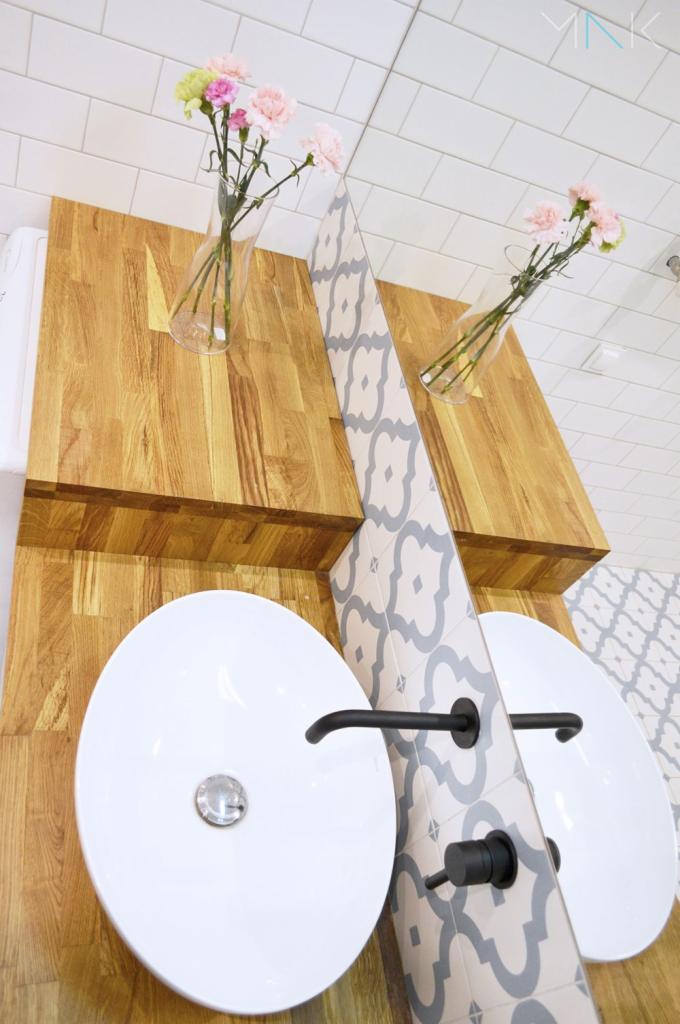 Dwie białe umywalki nablatowe z czarnymi kranami, osadzone na drewnianych blatach, z ozdobnymi wazonami z kwiatami i płytkami w tle.