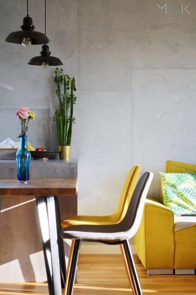 Nowoczesne wnętrze: drewniany stół z niebieskim wazonem i kwiatami, designerskie krzesła w odcieniach żółci i szarości, żółta sofa z poduszką w tropikalne liście, oświetlenie loftowe i szara ściana.