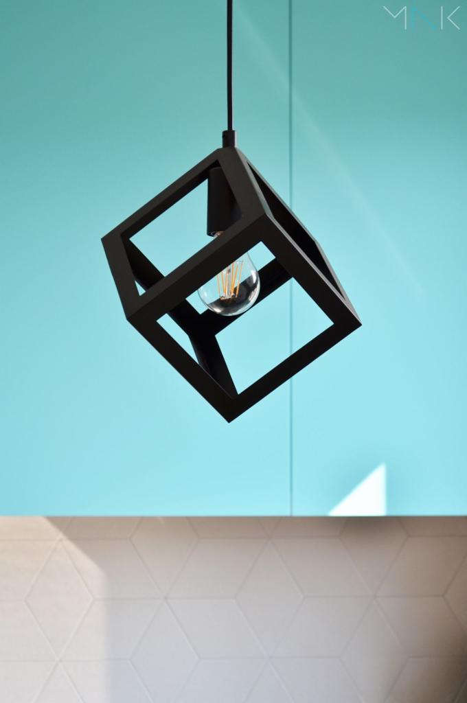 Czarna, geometryczna lampa wisząca w kształcie sześcianu z żarówką Edisona na tle turkusowej ściany i białych płytek z wzorem 3D.