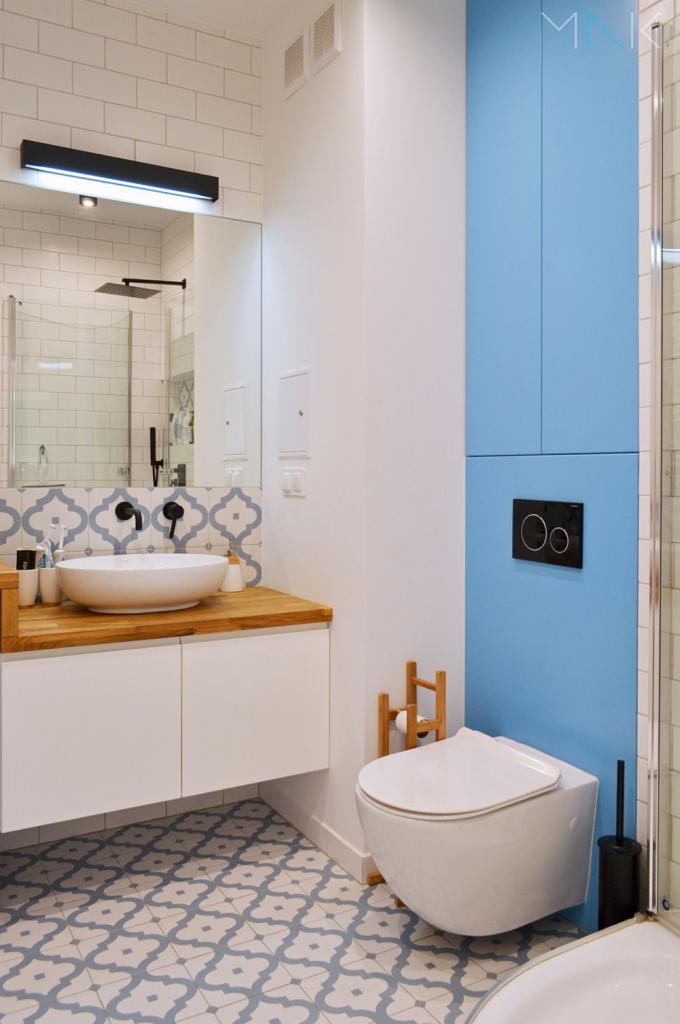 Nowoczesna łazienka z białą umywalką nablatową na drewnianym blacie, białą szafką, wiszącą toaletą na tle błękitnej ściany z szafkami, oraz wzorzystą podłogą w odcieniach szarości i błękitu.