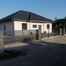 Nowoczesny dom jednorodzinny z szarą, poziomą bramą wjazdową i ogrodzeniem panelowym na froncie działki, widok w słoneczny dzień.