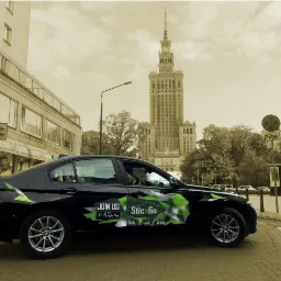 Czarny samochód osobowy z zielonymi elementami graficznymi i napisem 'SticnGo' na karoserii, zaparkowany na ulicy z widokiem na Pałac Kultury i Nauki w Warszawie.