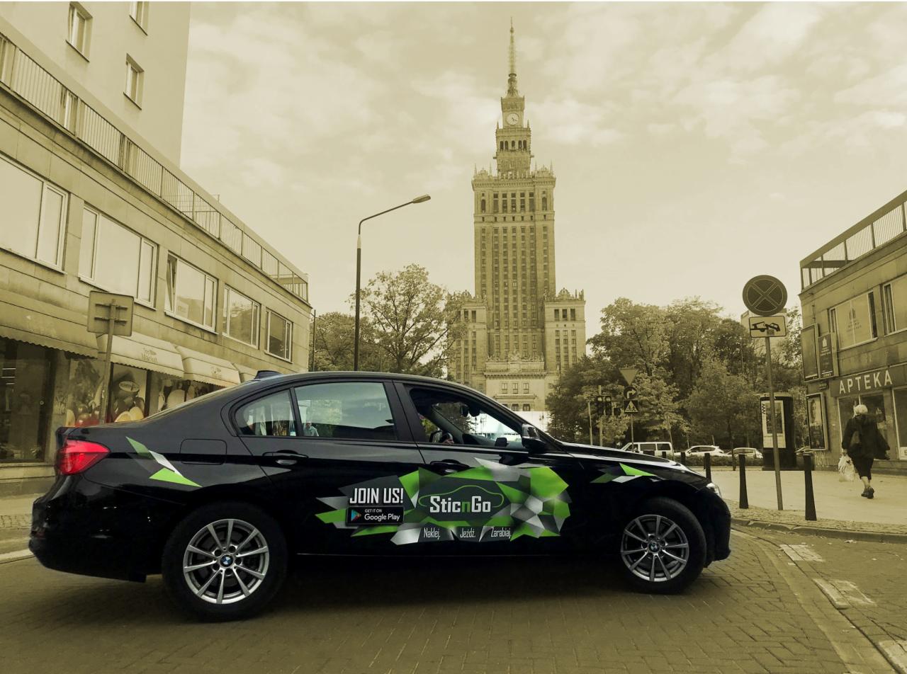 Czarny samochód osobowy z zielonymi elementami graficznymi i napisem 'SticnGo' na karoserii, zaparkowany na ulicy z widokiem na Pałac Kultury i Nauki w Warszawie.