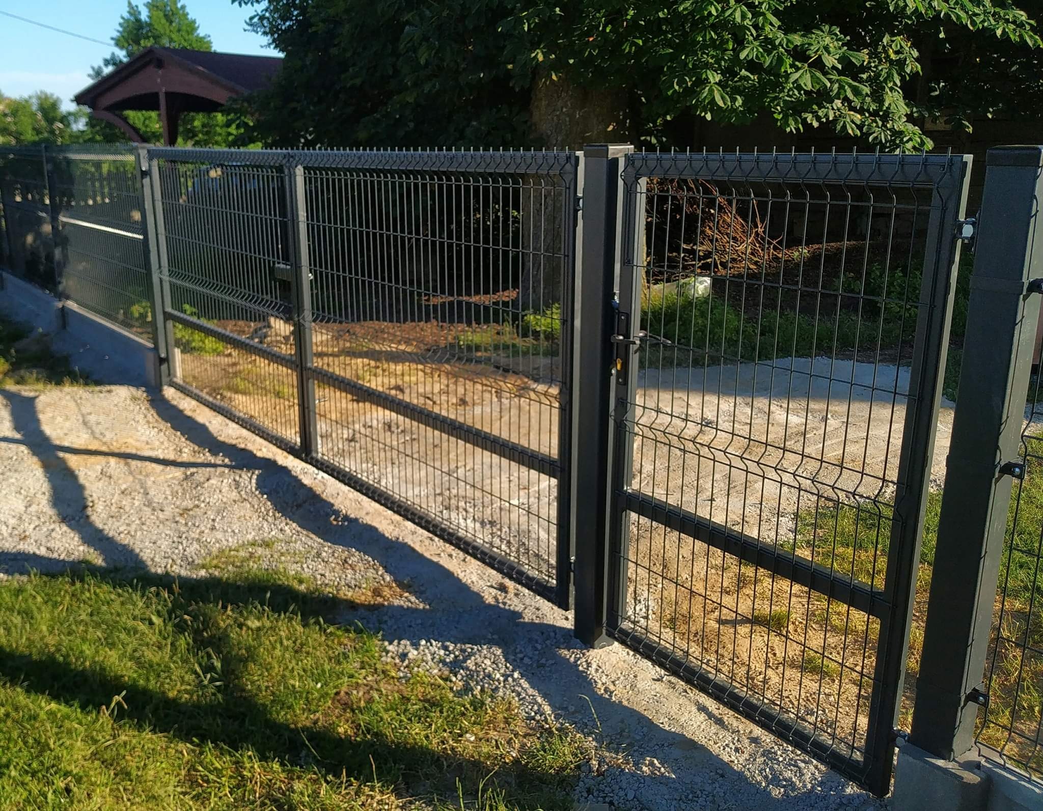 Nowoczesna, antracytowa brama panelowa z siatki z widocznym zamkiem, osadzona na betonowej podmurówce, otoczona trawnikiem i żwirową ścieżką, z zadaszeniem w tle.