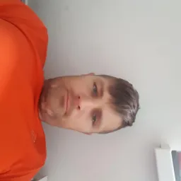 Mężczyzna w pomarańczowej koszulce sportowej, z krótką fryzurą, patrzy w obiektyw. Ujęcie z bliska, obrócone o 90 stopni w lewo.