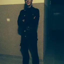 Uśmiechnięty mężczyzna w pełnym stroju roboczym, czapce z napisem i okularach, stoi na korytarzu z założonymi rękami.