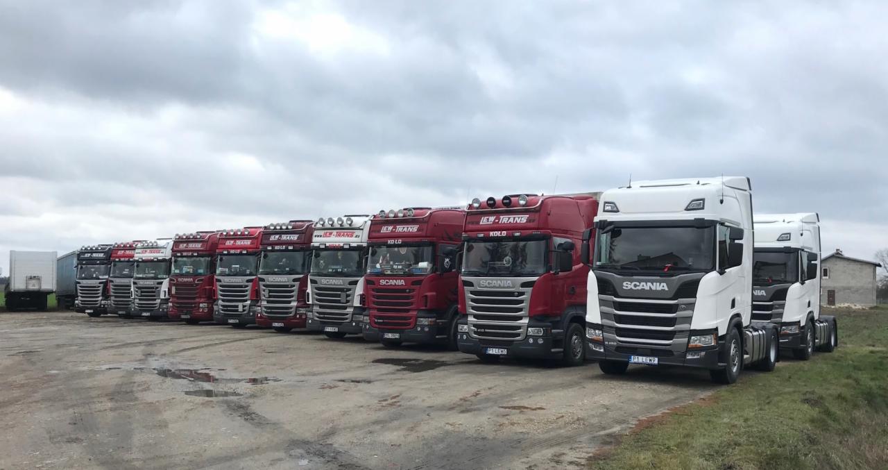 Flota ciężarówek Scania, w kolorach czerwonym i białym, zaparkowana na placu firmy transportowej, z widocznym budynkiem gospodarczym w tle.