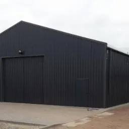 Gotowa, długa, ciemnoszara hala z blachy trapezowej z dwojgiem dużych wrót i małymi drzwiami z boku, oświetlona lampą zewnętrzną, na betonowej podbudowie.