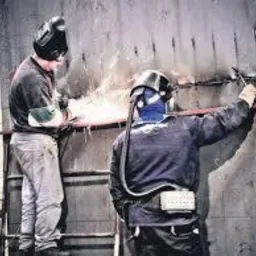 Spawanie pionowe blachy stalowej przez dwóch spawaczy w maskach ochronnych, iskry w powietrzu, widoczne spoiny i drabina.