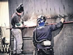 Spawanie pionowe blachy stalowej przez dwóch spawaczy w maskach ochronnych, iskry w powietrzu, widoczne spoiny i drabina.