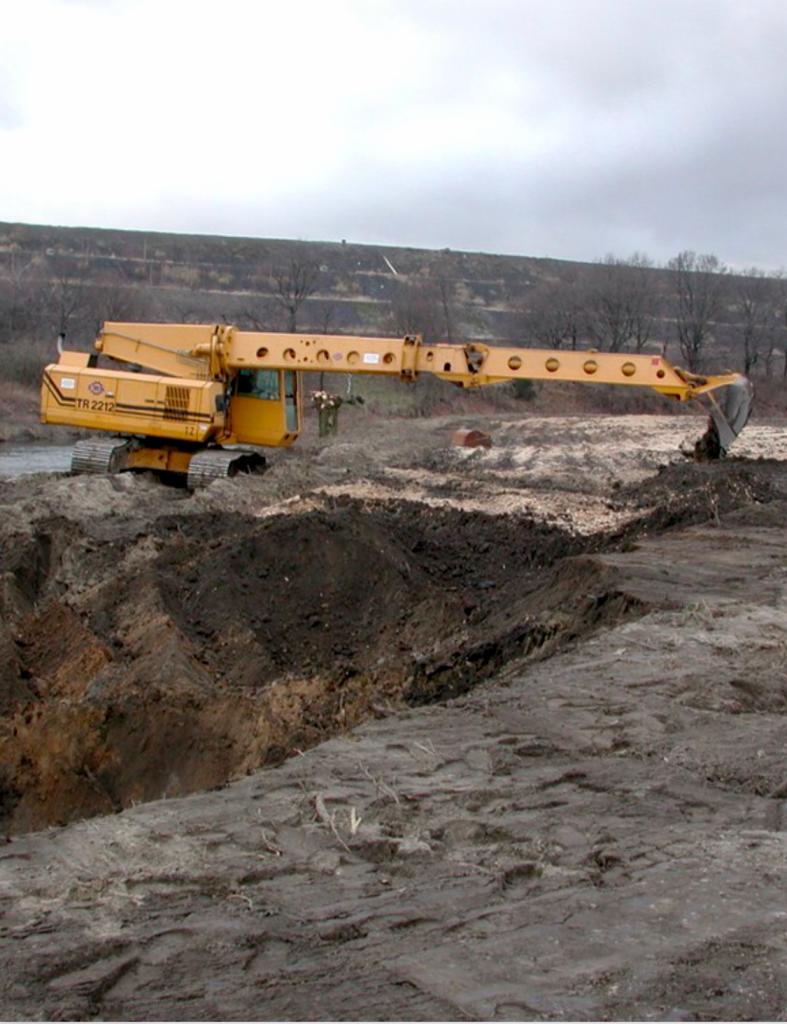 Żółta koparka gąsienicowa z długim wysięgnikiem pracująca przy wykopie ziemnym, w tle widać rzekę i pagórkowaty krajobraz pod pochmurnym niebem.