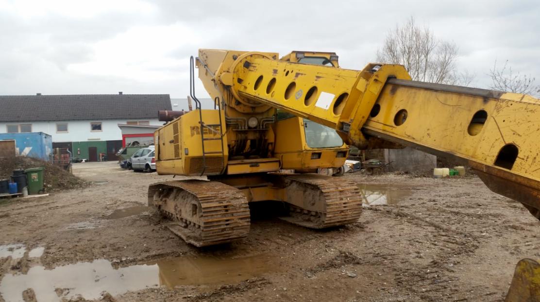 Żółta maszyna budowlana na gąsienicach, z długim wysięgnikiem, stoi na błotnistym terenie budowy z budynkami w tle