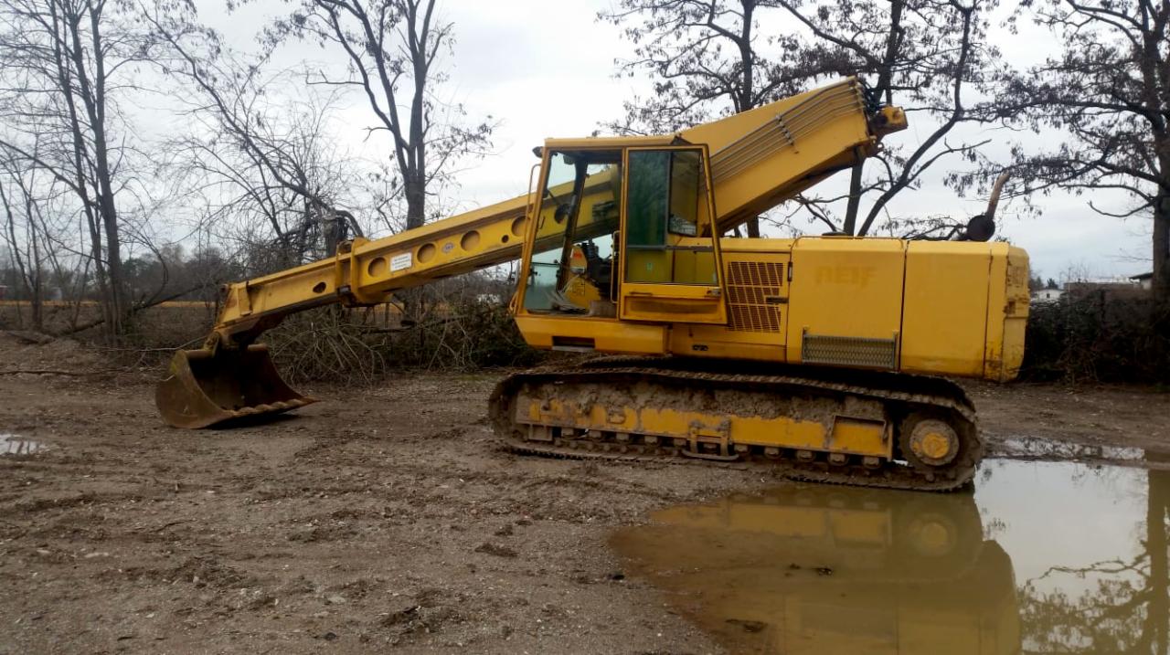 Żółta koparka gąsienicowa stoi na błotnistym terenie z kałużami, w tle widoczne drzewa bez liści i ścięte gałęzie.