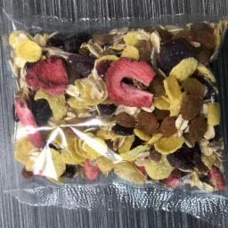 Przezroczyste opakowanie foliowe wypełnione mieszanką płatków zbożowych, suszonych owoców (truskawki, rodzynki, ciemne jagody) i ziaren, leżące na szarym tle.