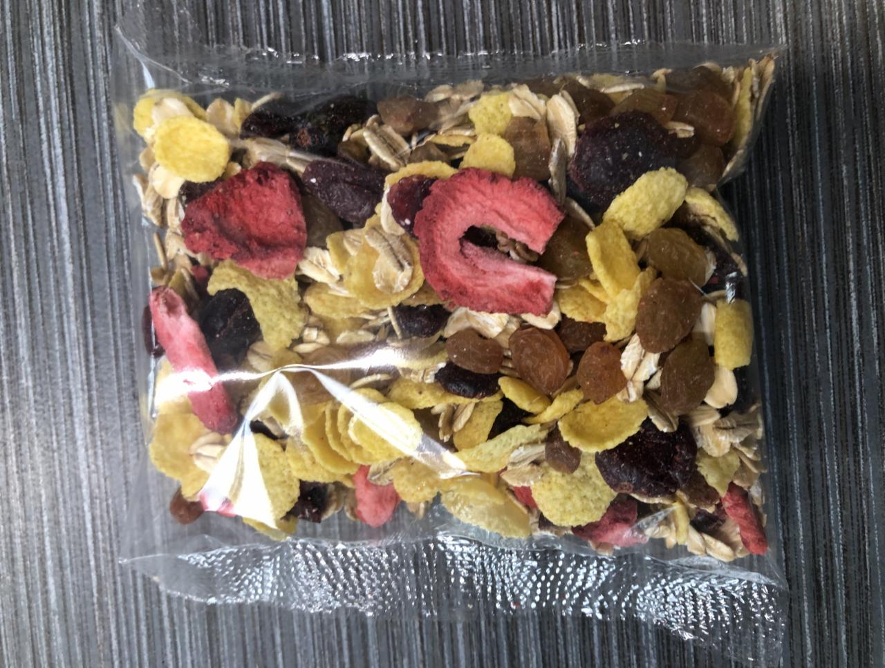 Przezroczyste opakowanie foliowe wypełnione mieszanką płatków zbożowych, suszonych owoców (truskawki, rodzynki, ciemne jagody) i ziaren, leżące na szarym tle.