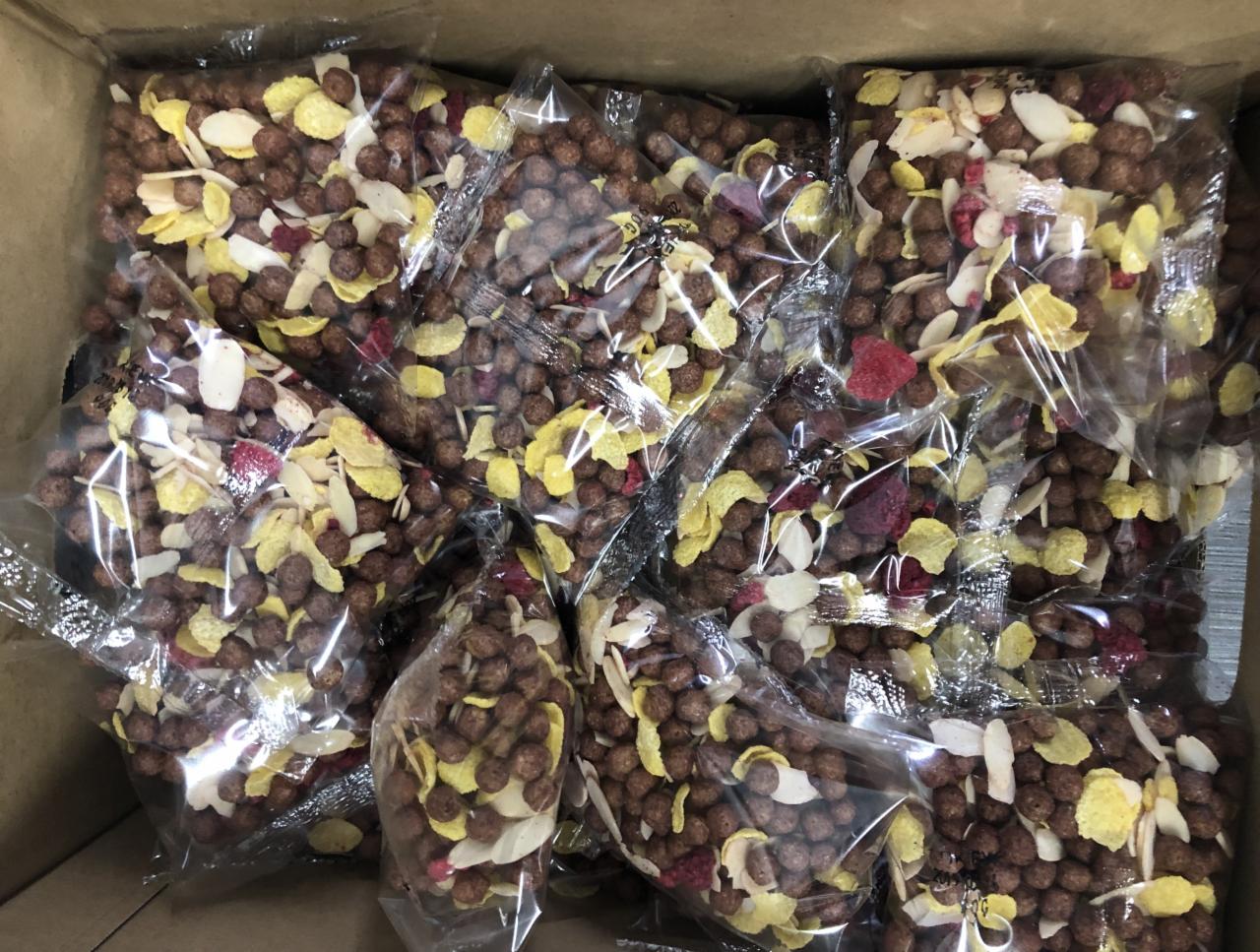 Pudełko wypełnione przezroczystymi woreczkami z brązowymi kulkami zbożowymi, płatkami kukurydzianymi i kawałkami suszonych owoców, widać nierównomierne wypełnienie woreczków.