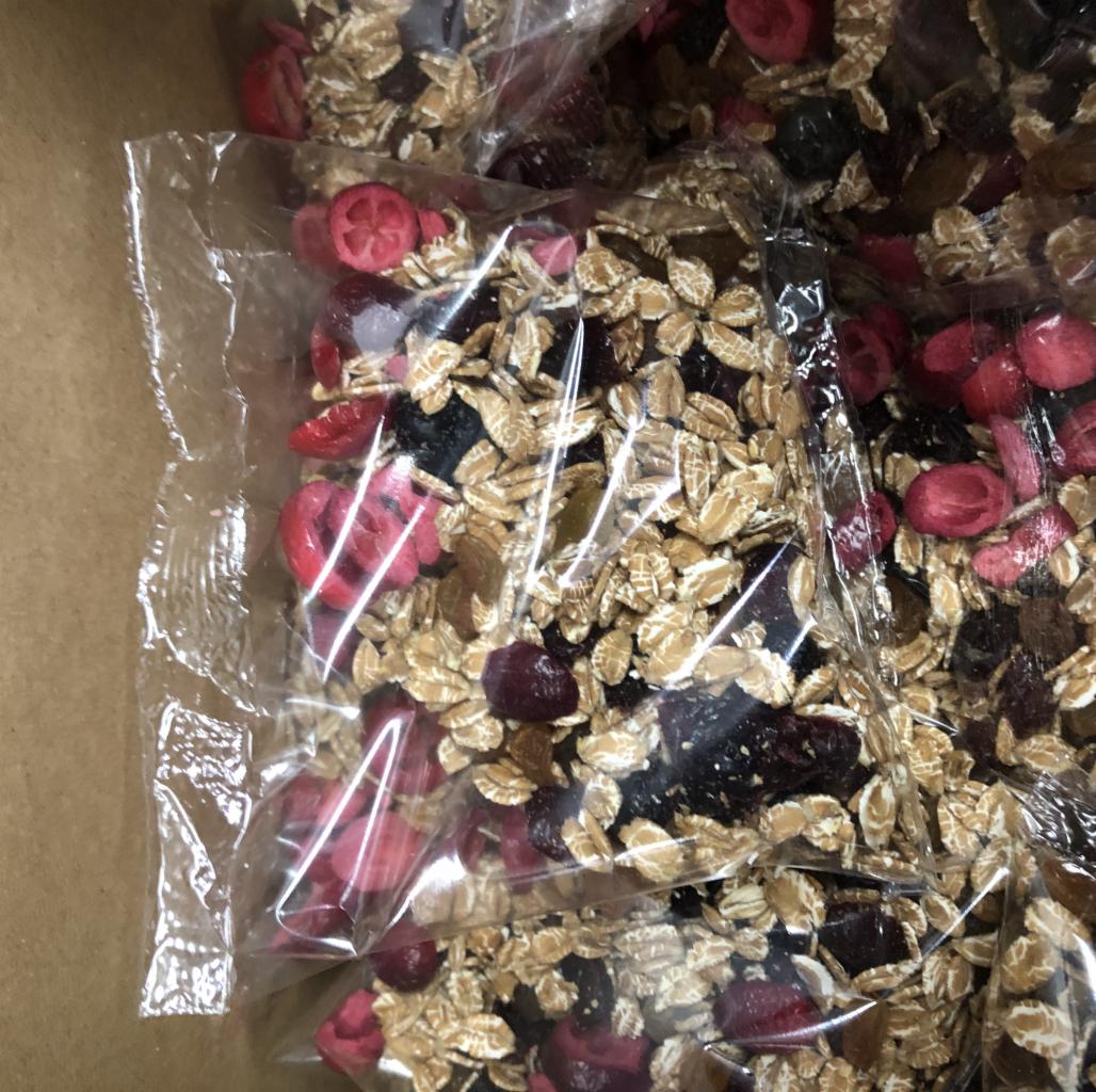 Pojedyncze porcje mieszanki płatków owsianych i suszonych owoców zapakowane w przezroczyste, foliowe woreczki, umieszczone w tekturowym pudełku.