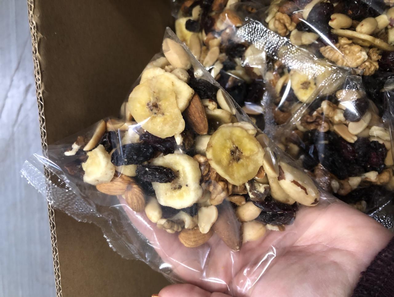 Dłoń trzyma przezroczysty woreczek foliowy wypełniony mieszanką orzechów, suszonych bananów i rodzynek, widoczne inne zapakowane porcje w kartonowym pudełku.