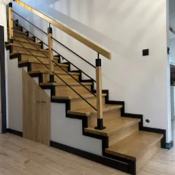 Nowoczesne schody drewniane z czarnymi elementami konstrukcyjnymi i metalową balustradą, zintegrowane ze schowkiem pod schodami, na tle białej ściany z widocznymi gniazdkami.