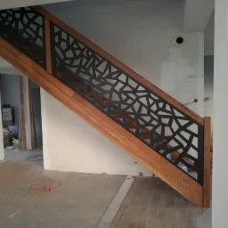 Drewniane schody z balustradą o geometrycznym, ażurowym wzorze w kolorze czarnym, widok z dołu, w tle surowy stan wykończenia wnętrza.