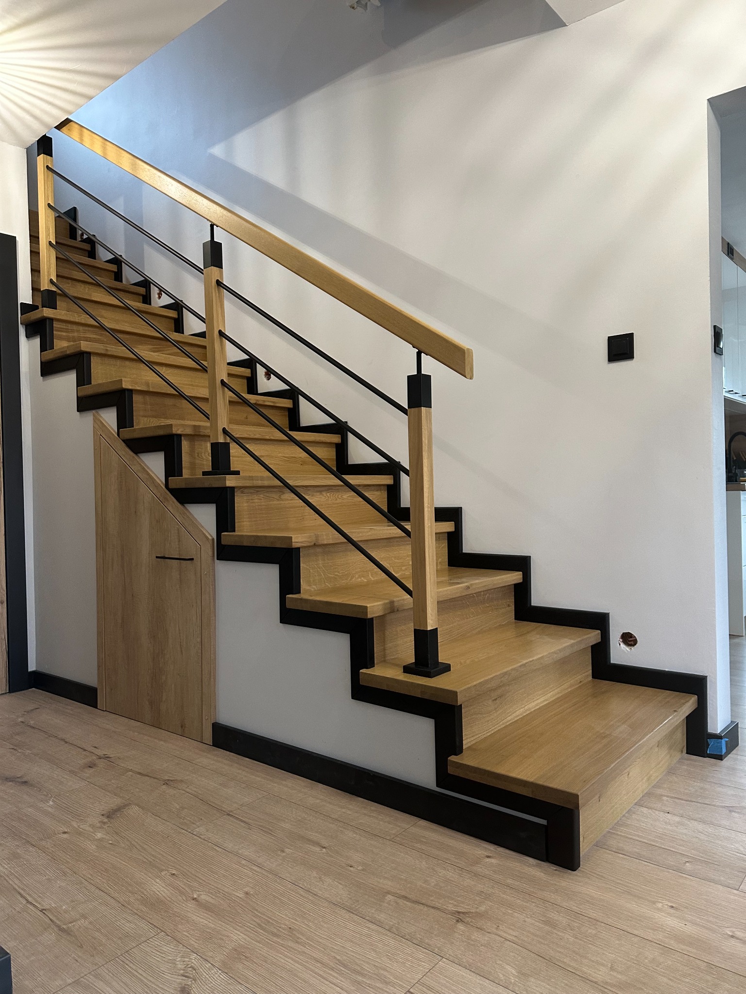 Nowoczesne schody drewniane z czarnymi elementami konstrukcyjnymi i metalową balustradą, zintegrowane ze schowkiem pod schodami, na tle białej ściany z widocznymi gniazdkami.